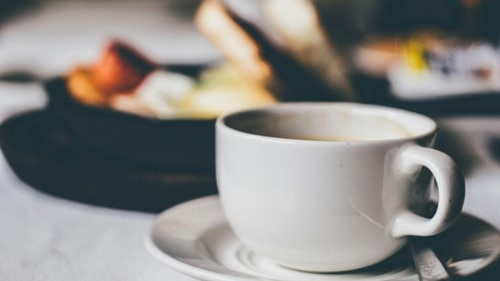 A half-drank white coffee mug placed on top of a white saucer next to a silver teaspoon. 