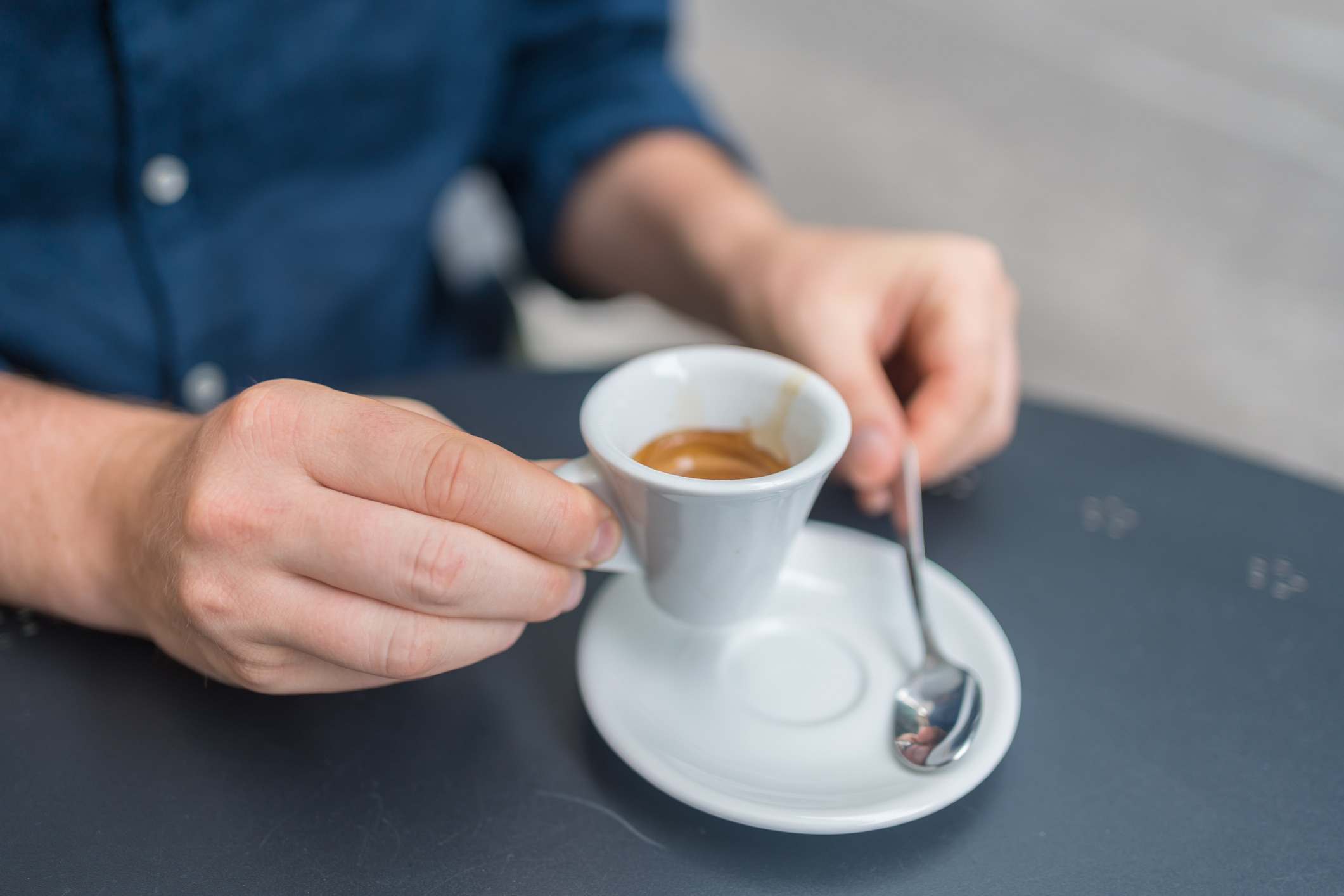 A person in a blue shirt holding a white espresso cup with a saucer and spoon on a dark table.